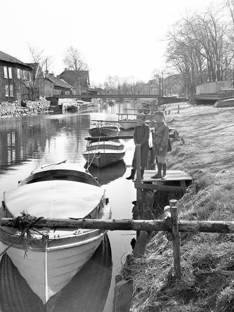 Båtar i Kungsbackaån. De två pojkarna verkar överväga att ta ett skutt ner i Kungsbackaån denna majdag år 1947. Deras klädsel utgör en fin tidstypisk kollektion: äppelknyckarbyxor, liten pirka på huvudet, reservylletröja, knähöga strumpor och gummistövlar. Gummistövlarna hade hört till den svenska standardrepertoaren för lekande barn sedan 1920-talet.   Kungsbackaån – längre upp kallad Lindomeån – har sitt utlopp i Kungsbackafjorden. En antydan om Kungsbackaåns besvärande benägenhet att översvämmas i samband med snösmältningen anas i de höga stenskodda husgrunderna till vänster. 